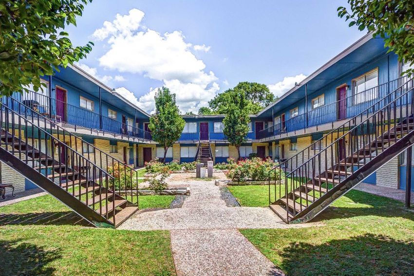 A courtyard view of a two-story apartment complex, featuring multiple units with blue siding and red doors. The area is landscaped with green grass, trees, and flower beds. Staircases lead to the second floor, and the sky is bright with fluffy clouds.