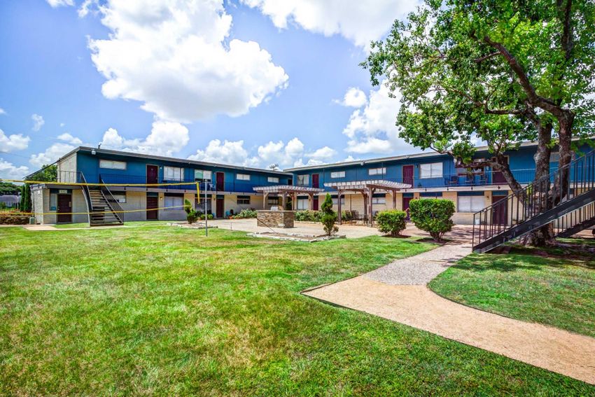 A multi-unit residential building with two levels, featuring a central courtyard with landscaped grass and shrubs. The building has a staircase on one side and a pathway leading through the grass. The sky is partly cloudy, adding to the bright atmosphere of the area.