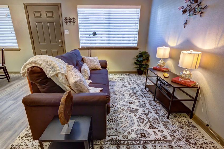 A cozy living room featuring a brown sofa with decorative pillows, a coffee table, and a lighted floor lamp. The room has two warm lamps on a side table, a leafy plant, and a patterned area rug. Natural light filters through window blinds, enhancing the inviting atmosphere.
