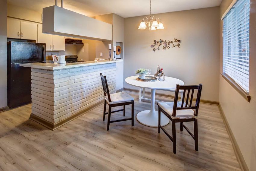 A modern kitchen and dining area featuring a round white table with two black chairs, a light-colored wooden floor, and a kitchen counter made of stone. The kitchen includes a black refrigerator and a range hood, with overhead lighting and decorative wall art in the background. Natural light streams in through a window.