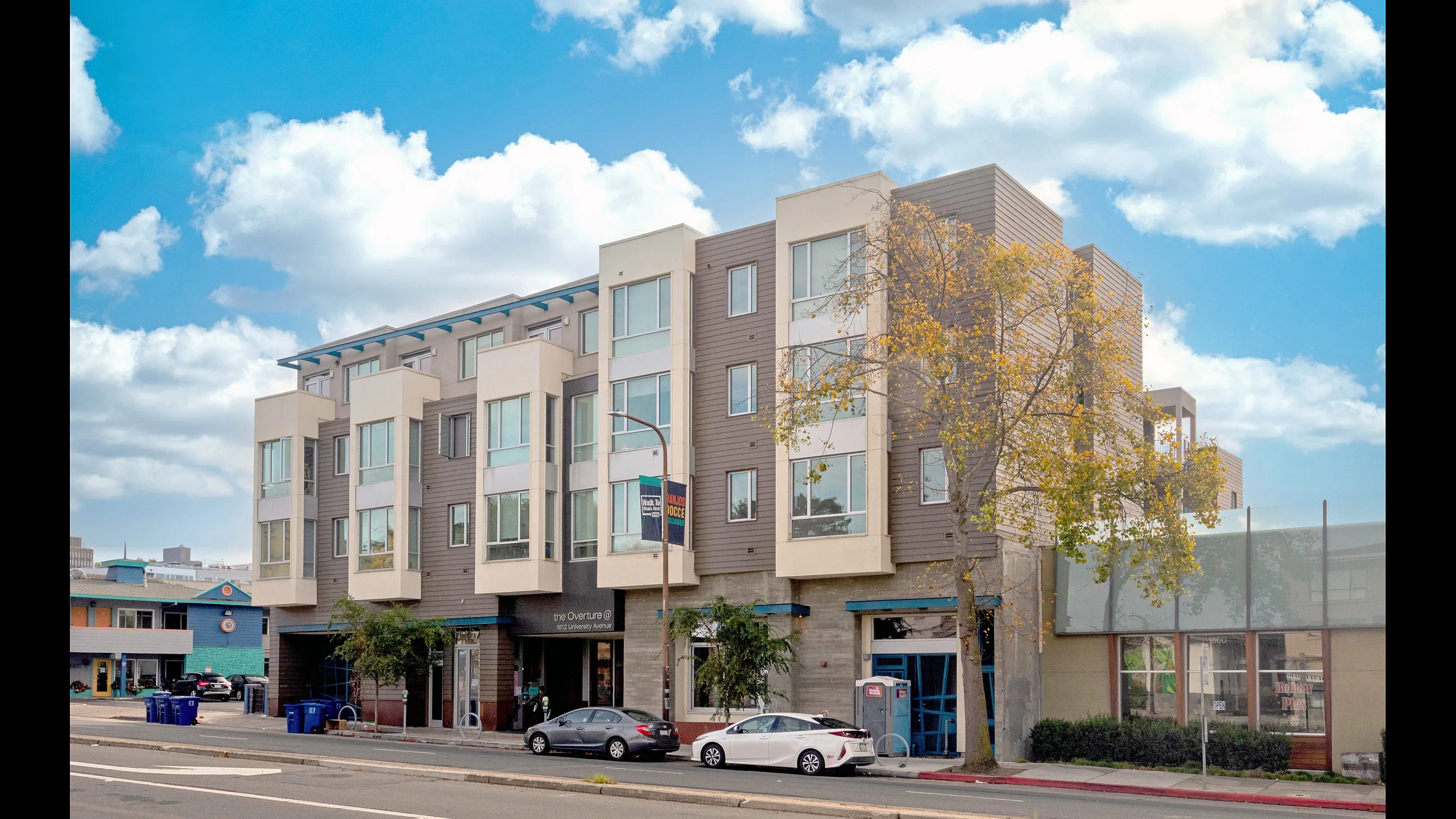 Modern urban apartment building with large windows and tree-lined street, set against a bright blue sky with fluffy clouds.