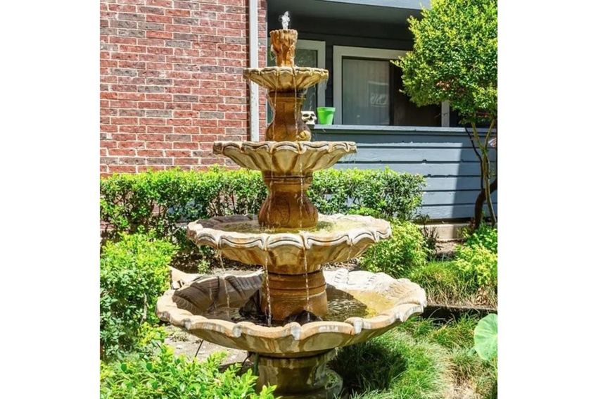 A multi-tiered stone fountain surrounded by lush greenery and small shrubs. The fountain has water flowing from the top tier down to the lower levels, creating a soothing water feature in a garden setting. A brick building is visible in the background.