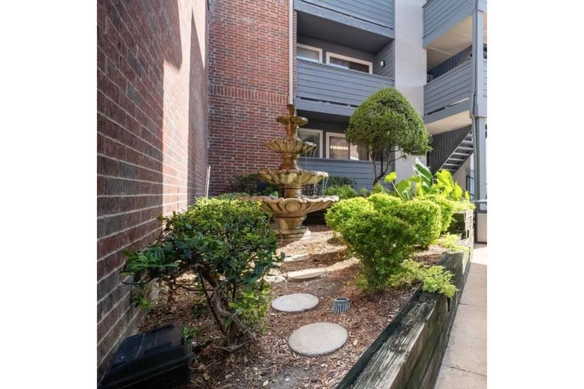 A landscaped area featuring a decorative stone fountain surrounded by neatly trimmed bushes and plants. The setting includes a mix of greenery and stone paths, with an adjacent building visible in the background. The overall atmosphere is calm and inviting.
