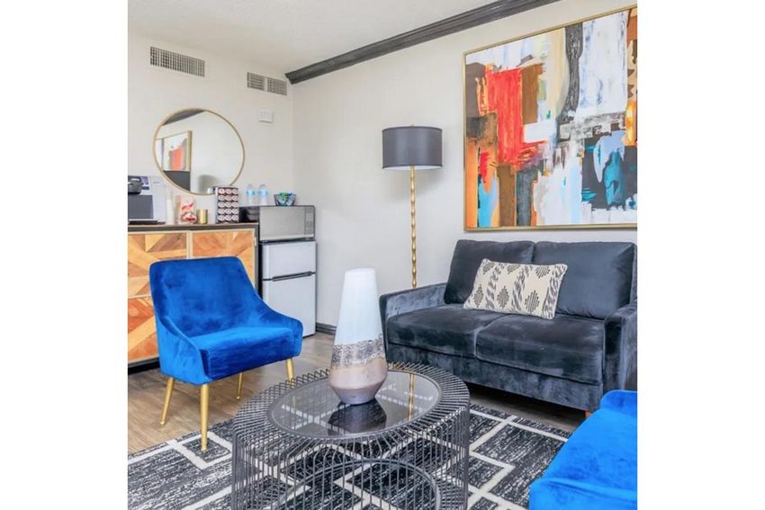 A stylish living room featuring a dark gray sofa, two blue chairs, and a round black coffee table. A decorative lamp stands beside the sofa. The wall art is colorful and abstract. In the background, there's a cabinet with drinks and a mirrored circle on the wall, enhancing the modern decor.
