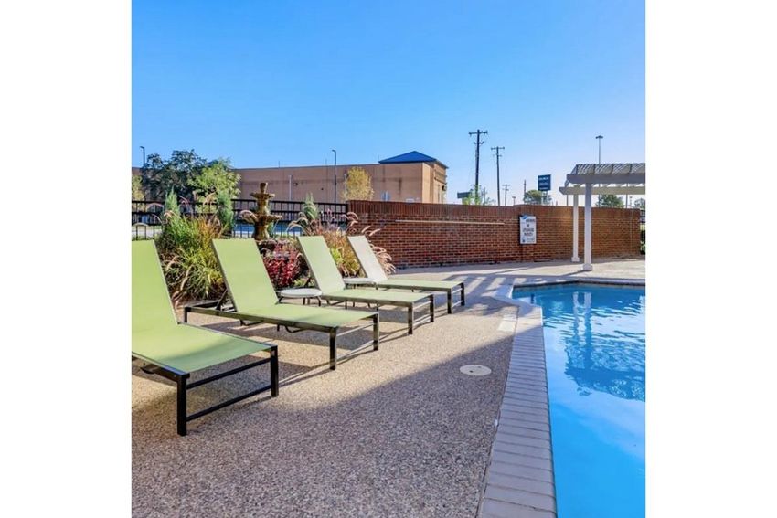 A serene outdoor area featuring a swimming pool bordered by a light tan patio. Several green lounge chairs are arranged beside the pool. In the background, there are brick walls and plants, with a building visible in the distance under a clear blue sky.