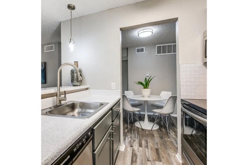 Modern kitchen featuring a stainless steel sink, sleek cabinetry, and a countertop with a light speckled design. In the background, a dining area with white chairs and a round table is visible. Soft lighting and neutral colors create a bright, inviting atmosphere.