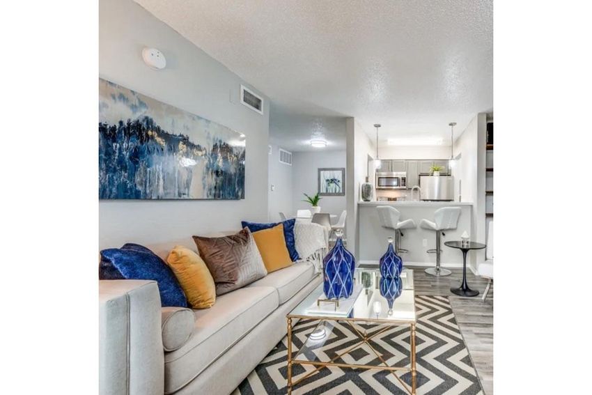 A modern living space featuring a light gray sofa adorned with colorful pillows, a stylish coffee table with decorative items, and abstract wall art. The room has an open layout leading to a kitchen with bar seating, complemented by neutral tones and vibrant accents in decor.
