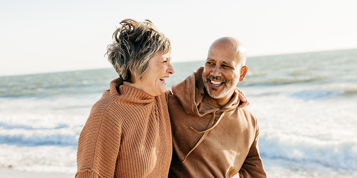 Joyful couple enjoying a sunny day at the beach, wrapped in cozy sweaters, with waves gently crashing in the background.