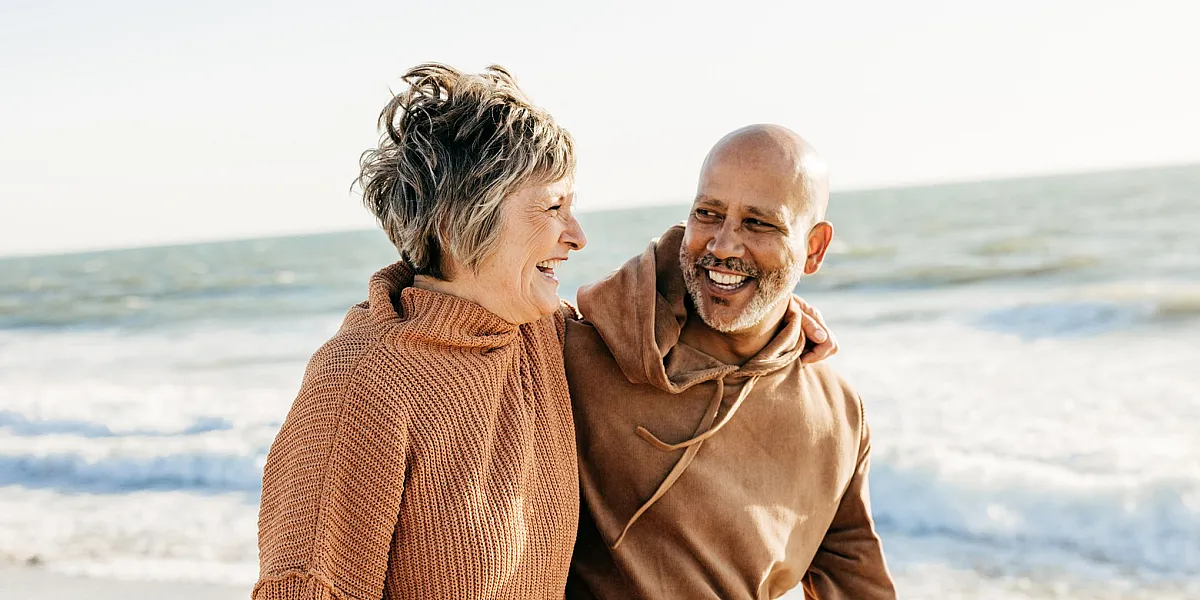 Joyful couple enjoying a sunny day at the beach, wrapped in cozy sweaters, with waves gently crashing in the background.