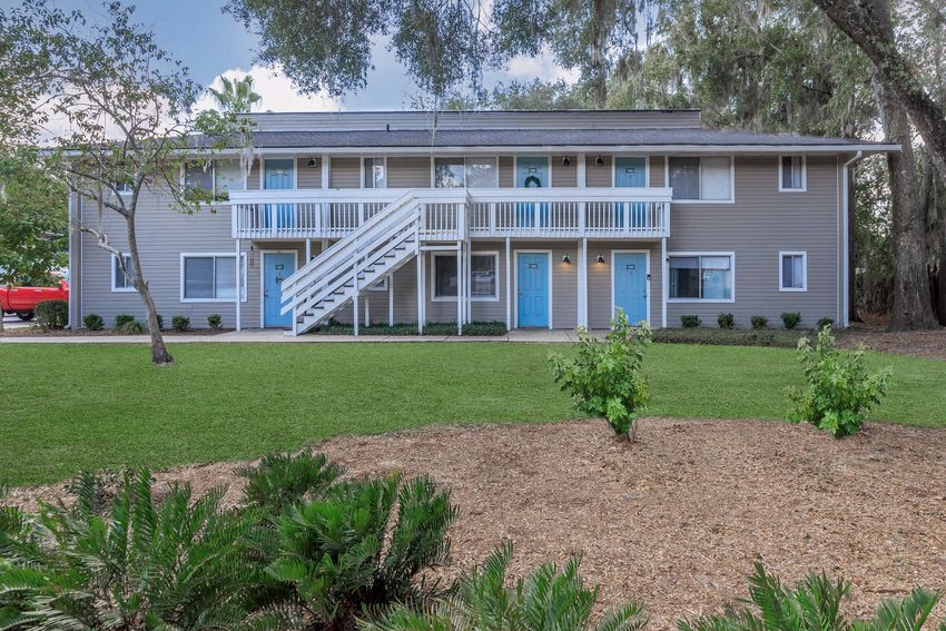 A two-story residential building with light gray siding and blue doors. The structure features two sets of stairs leading to the second floor, surrounded by a well-maintained lawn with shrubs and small trees. The setting is peaceful with green grass and decorative landscaping.