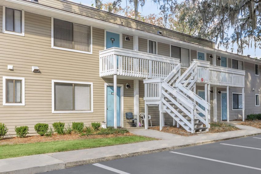 Two-story apartment building with a light brown exterior and white trim. The building features blue doors, a white wooden staircase leading to the second floor, and neatly trimmed shrubs along the front. Several parking spaces are visible in front of the building, set in a suburban environment with grass and trees.