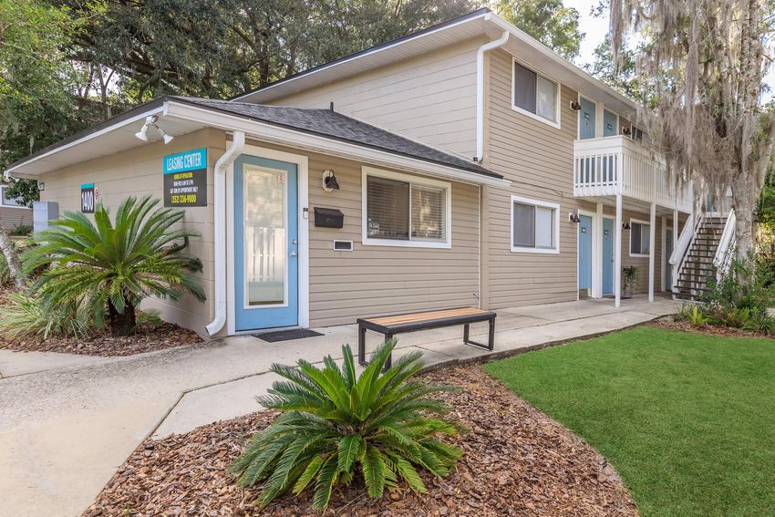 A two-story, multi-unit residential building with a light gray exterior and a blue front door. The entrance features a small bench and a landscaped area with ornamental plants. There are stairs leading to the upper unit, and the surroundings include green grass and mulch.