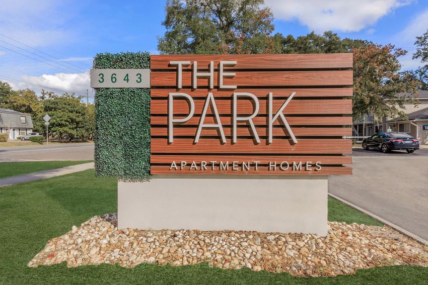 Sign for "The Park Apartment Homes" featuring modern wooden slats with green accents. The address "3643" is displayed on the left. The sign is surrounded by decorative stones and grass, with a clear blue sky in the background.