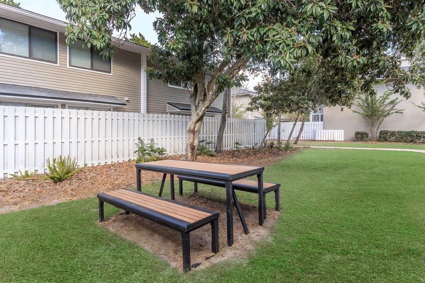 A picnic table made of wood and metal located on a grassy area, surrounded by trees and a white picket fence. The background features residential buildings, creating a serene outdoor setting suitable for gatherings or relaxation.