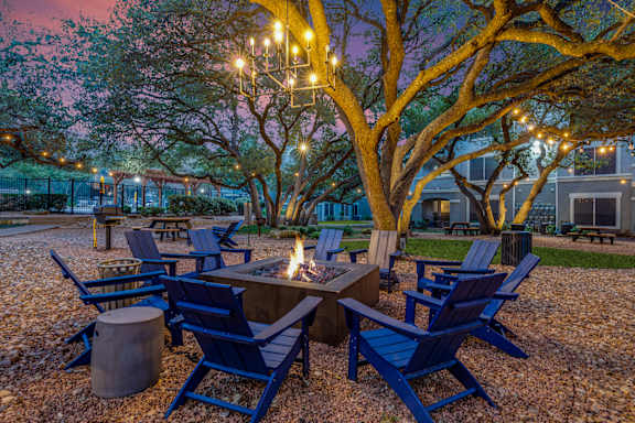 Outdoor community courtyard with Adirondack chairs arranged around a central fire pit, illuminated by string lights and a chandelier beneath mature trees at dusk.