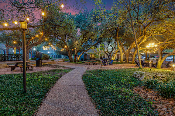 Tree-lined community courtyard with a winding concrete walkway, picnic tables, and lounge seating, softly lit by string lights at dusk.