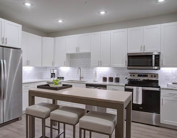 A modern kitchen with a stainless steel refrigerator and a central island with a cutting board and four stools.