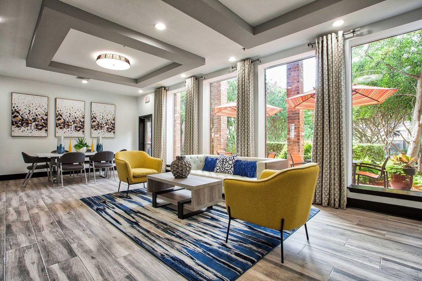 A modern living area features a yellow sofa and armchair, a coffee table, and a decorative vase. Large windows provide natural light and views of an outdoor patio with umbrellas. Light wood flooring complements the room, which is adorned with abstract wall art and a dining space in the background.