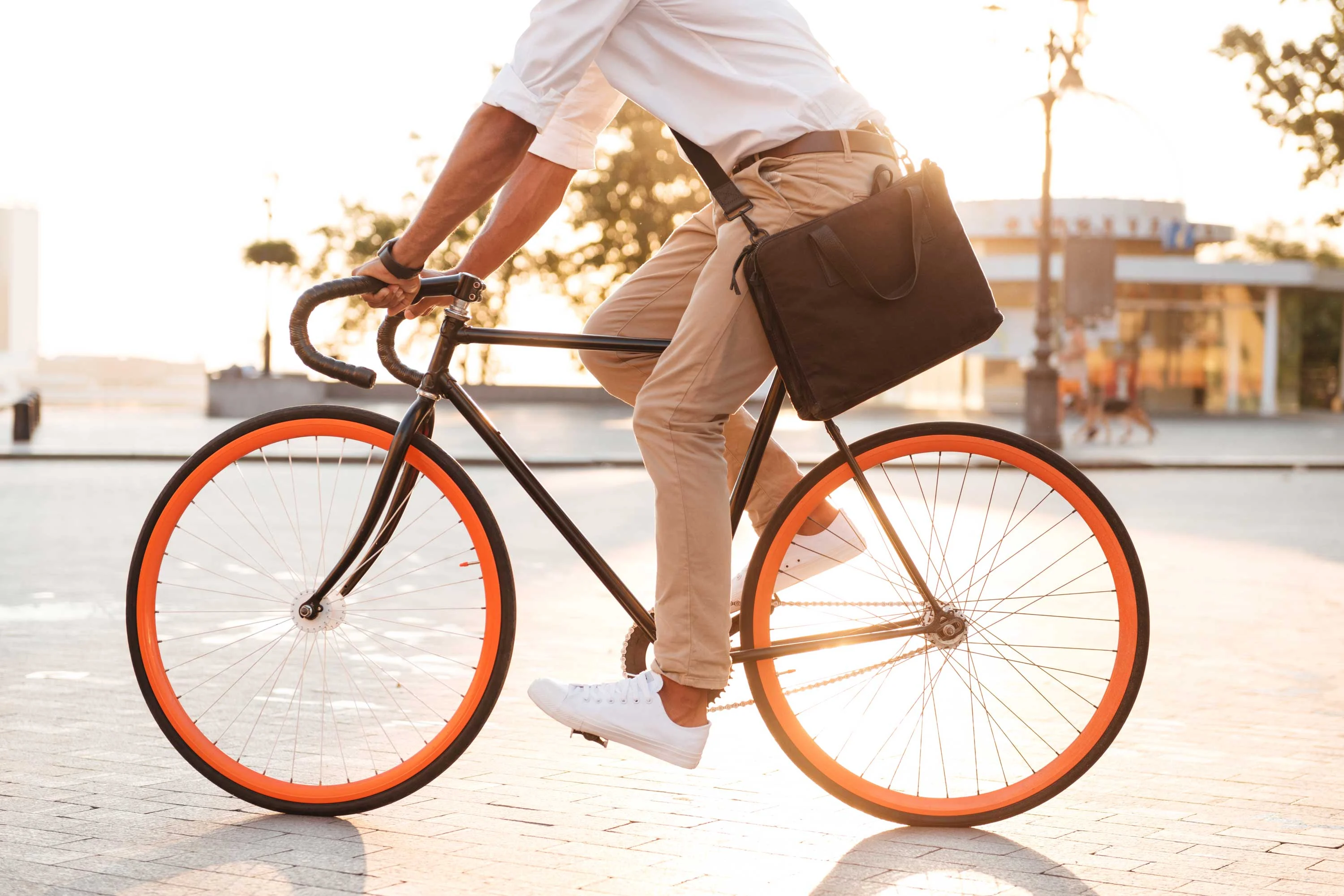 A man riding a bicycle with orange wheels in a sunlit outdoor setting, wearing a white shirt and beige pants, and carrying a black messenger bag.