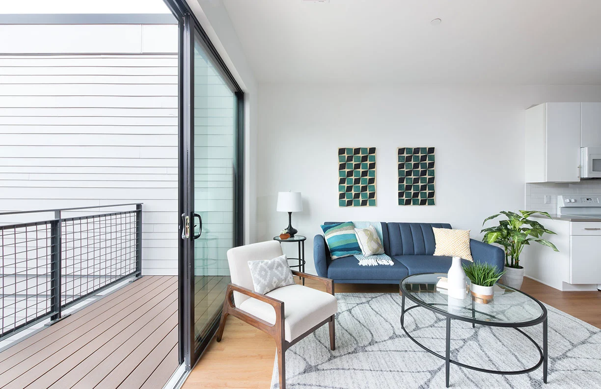 A modern living room featuring a blue sofa, a glass coffee table, and a view of a balcony through large sliding doors.