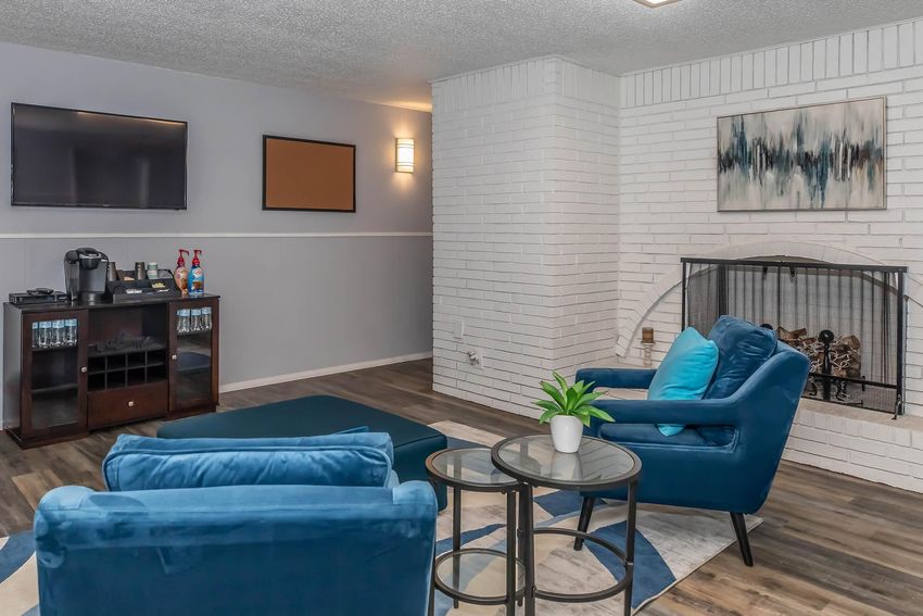 A cozy living room featuring two teal chairs, a small round table, and a modern fireplace. The walls are painted in light gray and white, with a flat-screen TV mounted on one wall. There is a decorative painting above the fireplace and a coffee station in the corner. Natural light enhances the inviting atmosphere.