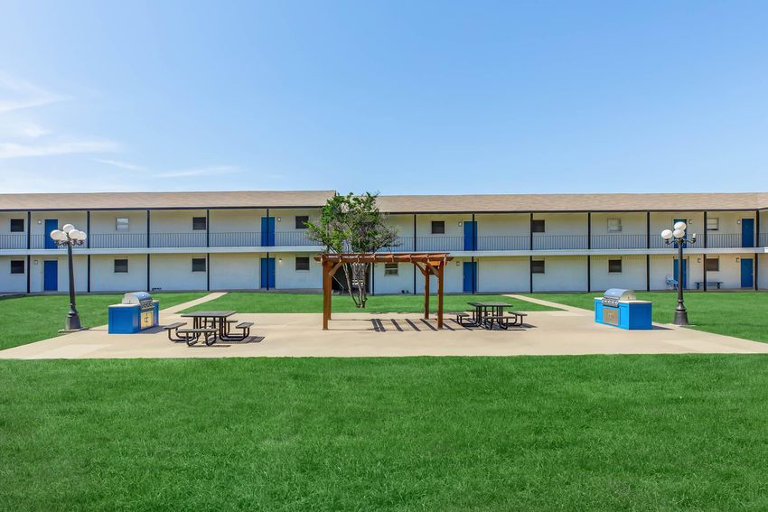 A courtyard area featuring a picnic table set under a wooden gazebo, surrounded by green grass. In the background, there are two-story buildings with blue doors and light-colored walls. Two trash bins are positioned near the picnic area. The sky is clear and blue.