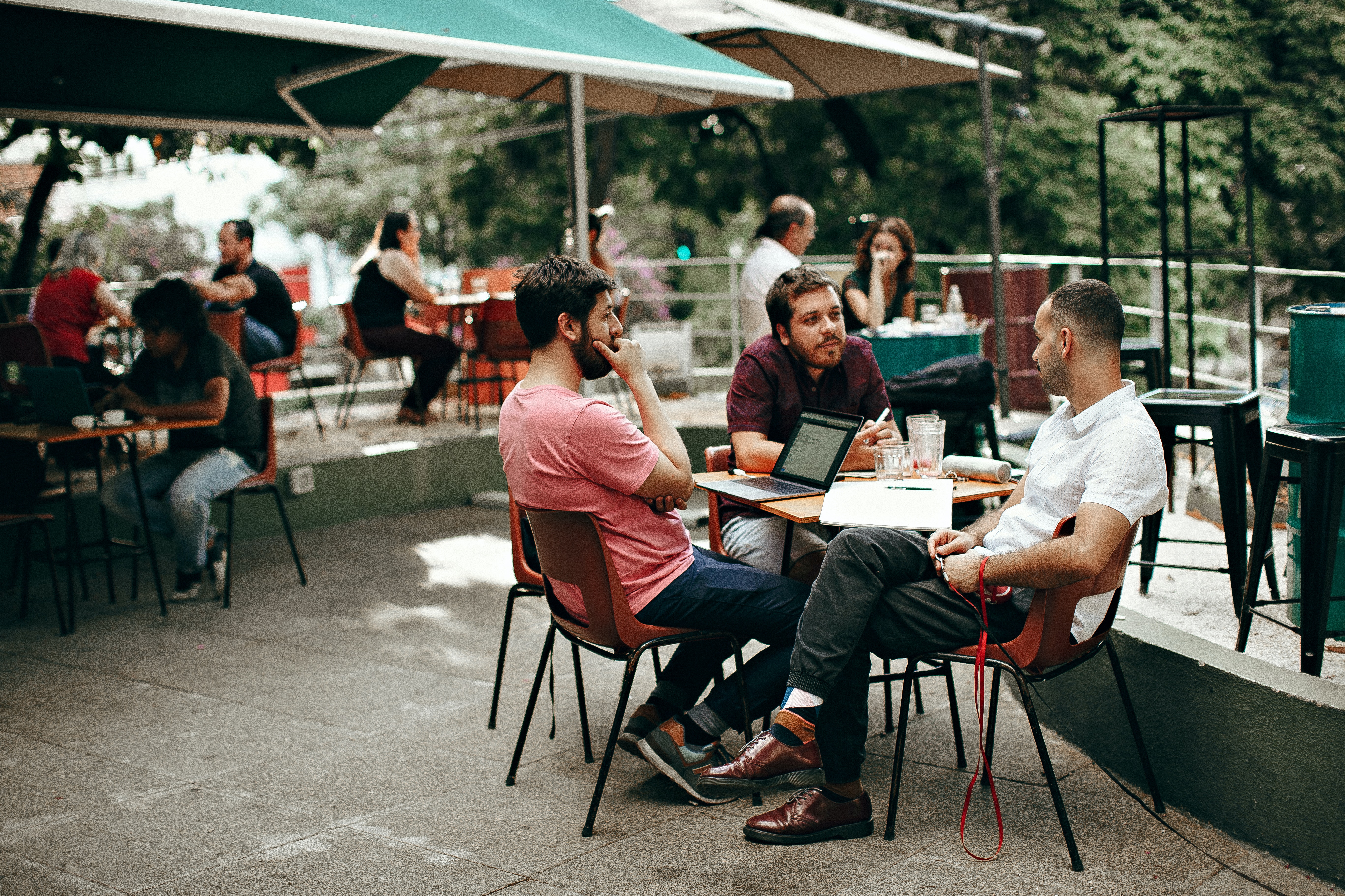 A group of three men engaged in conversation at an outdoor café, with tables and patrons in the background.