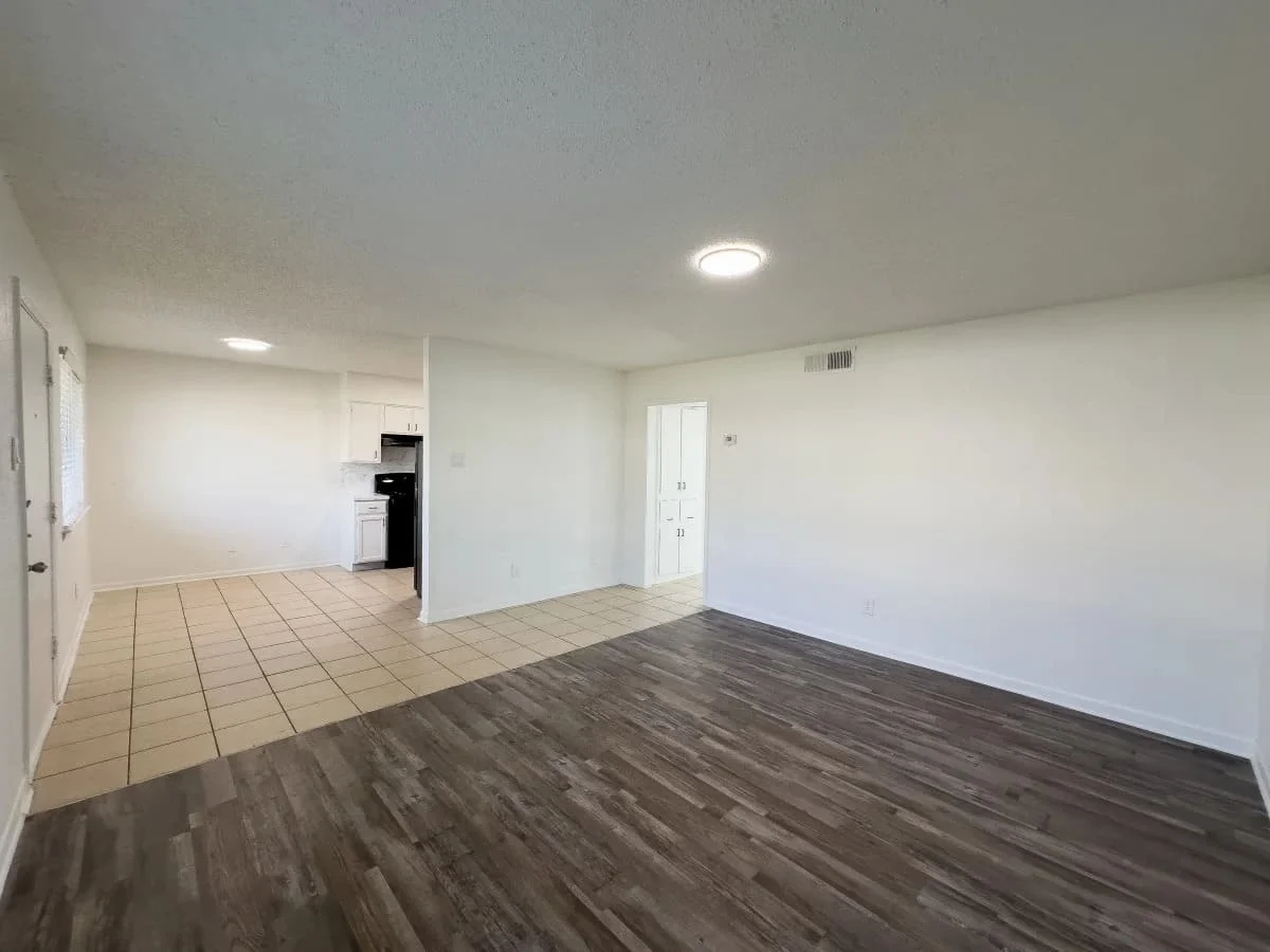 A spacious living area featuring a combination of tiled and wooden flooring. On the left, there is a doorway leading to a kitchen with black appliances. The walls are painted white, and there is overhead lighting.
