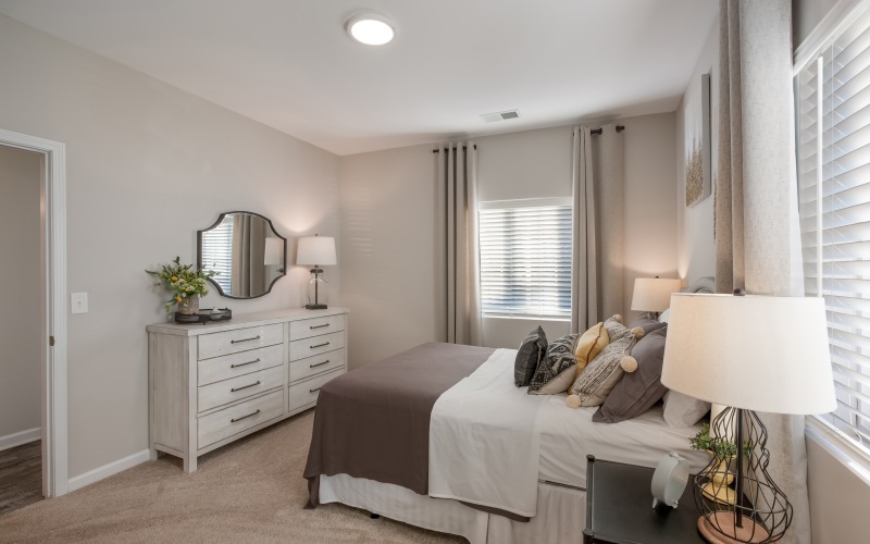 An interior view of a bedroom with a bed, chest of drawers, a mirror, side tables with lamps, and windows with curtains at The Summit on 401 pet friendly apartments in Fayetteville, NC.