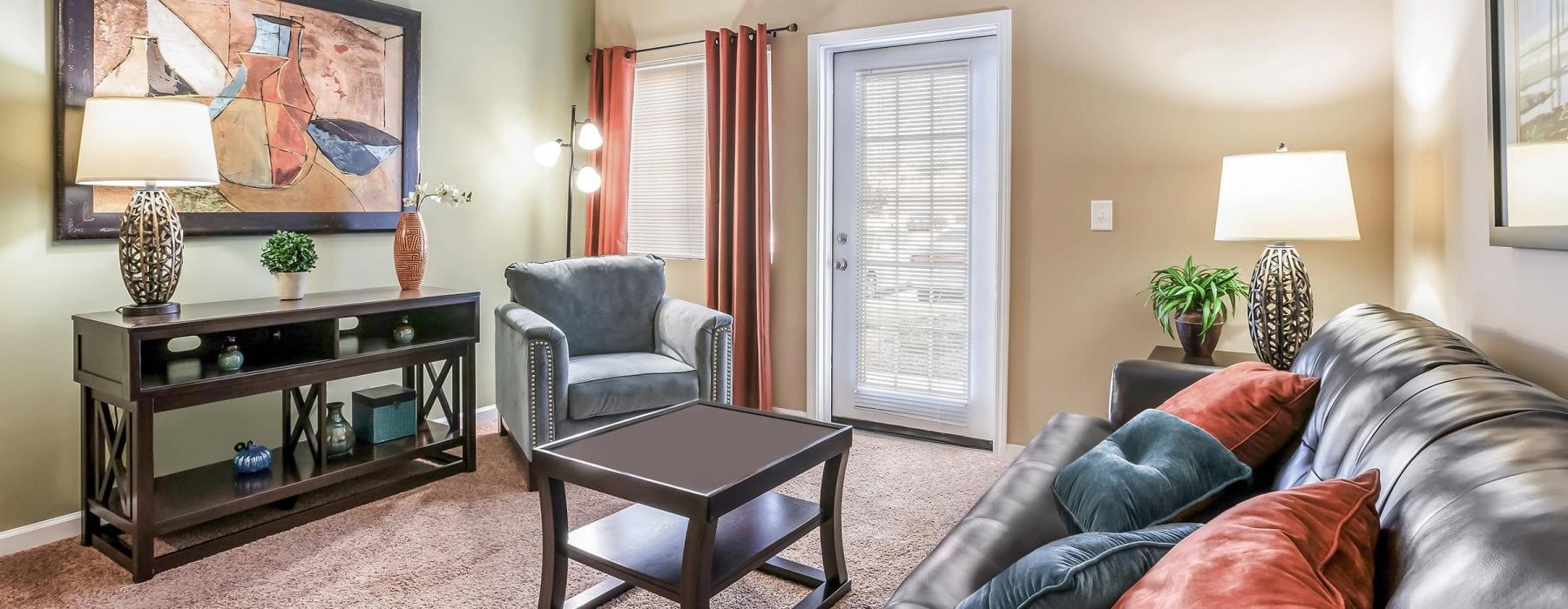 An interior view of a living room with a couch, a coffee table, a chair, a window with curtains, and a patio door at The Summit on 401 apartments for rent in Fayetteville, North Carolina.