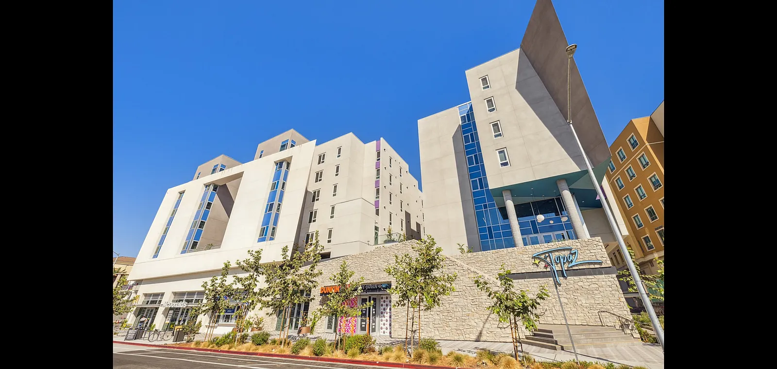 Modern apartment building with sleek architecture, featuring large windows and vibrant retail stores at street level under a clear blue sky.