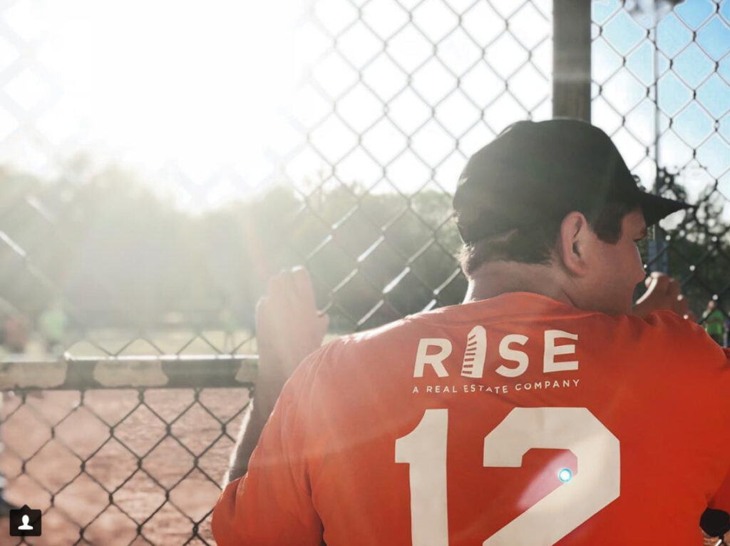 Man in orange RISE shirt with number 12, at a baseball field fence, sunlight in the background.
