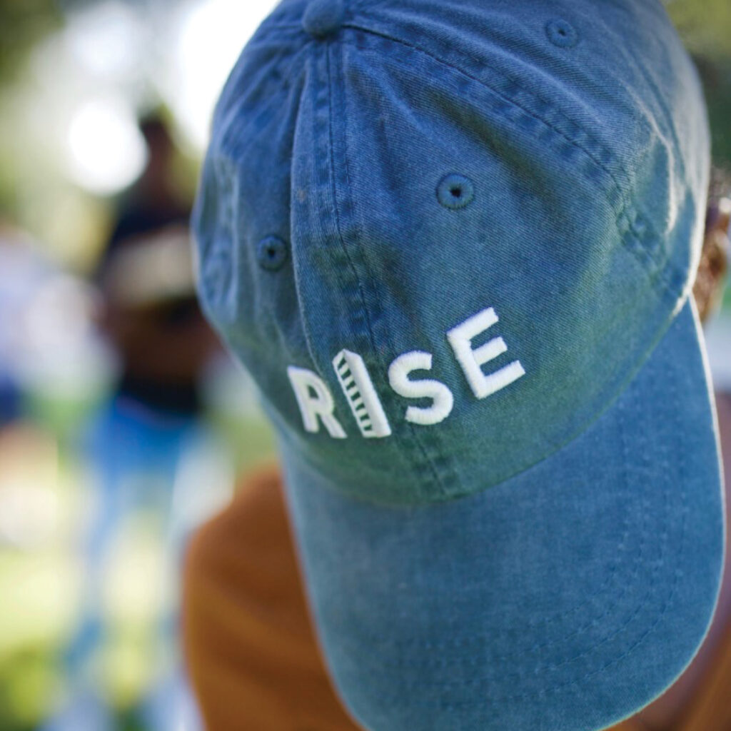 Blue baseball cap with RISE embroidery in white, worn outdoors, blurred background of people and greenery.