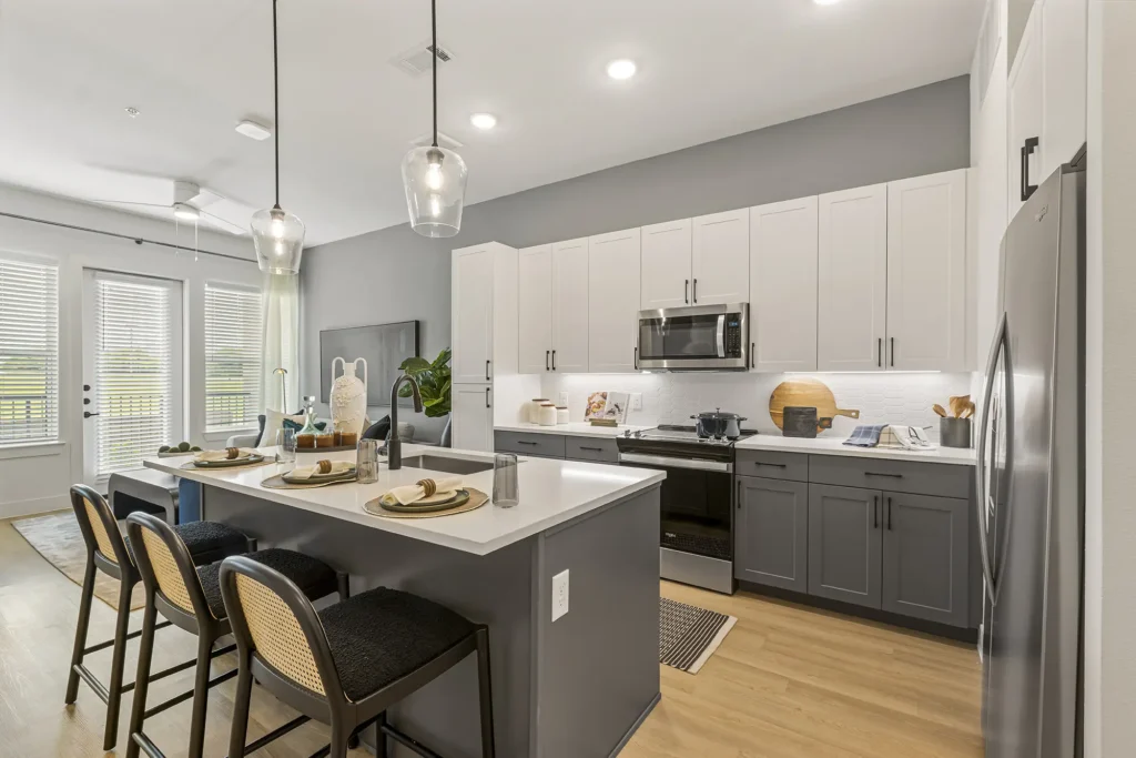 Modern kitchen with white upper cabinets and gray lower cabinets, stainless steel appliances, a white island with three barstools, pendant lights, and a dining setup. Large windows provide ample natural light.