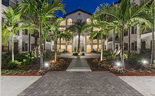 A pathway leads to a building with a lit entrance, flanked by palm trees and shrubbery.