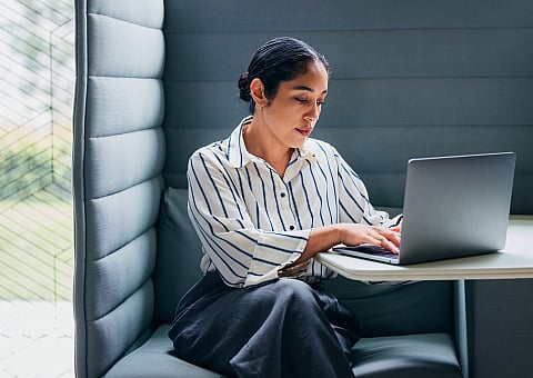 Focused professional woman working on a laptop in a cozy, modern booth with ambient lighting.