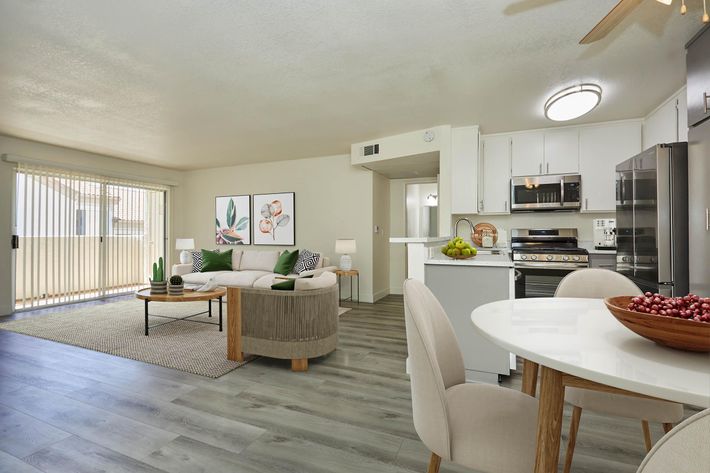 Bright and modern living space featuring a comfortable sofa, dining area with a wooden table and fruit bowl, and sleek kitchen appliances. Large windows allow natural light and open to a balcony. Decorative plants and artwork add a touch of style to the cozy atmosphere.