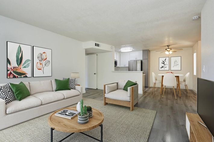 A modern living room featuring a light-colored sofa with green and black accents, a wooden coffee table with decor, and two framed botanical prints on the wall. The space transitions to a dining area with a light wood table and white chairs, illuminated by a ceiling fan, creating an inviting atmosphere.
