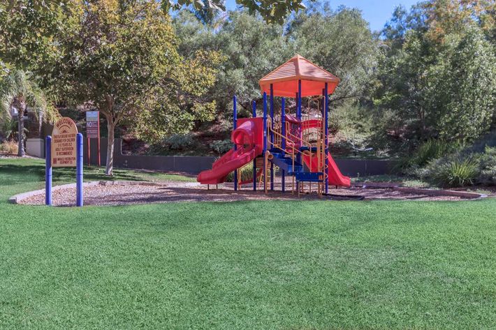 A brightly colored playground structure with slides and climbing features, situated on lush green grass surrounded by trees and landscaping.