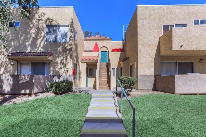 Two multi-unit buildings with beige exteriors, a greenish-blue door at the top of a staircase, and a red umbrella visible above. Lush green grass surrounds the pathway leading to the entrance, and there are some trees casting shadows.