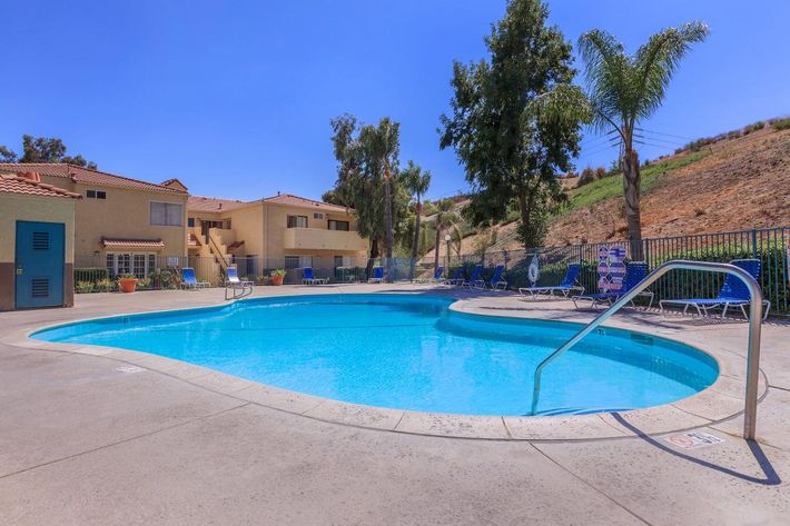 A clear blue swimming pool surrounded by lounge chairs, palm trees, and residential buildings in the background on a sunny day.