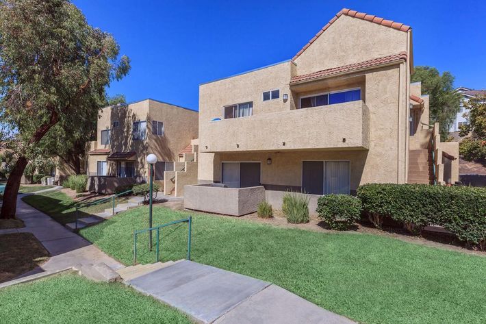 Two residential buildings with beige stucco exteriors, featuring balconies and landscaped lawns. The foreground includes a paved walkway with a railing, and the background shows a clear blue sky.