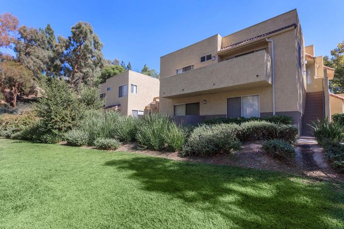 Two residential buildings with a light-colored exterior, situated beside a well-maintained green lawn. The landscape features shrubs and trees in the background, under a clear blue sky.