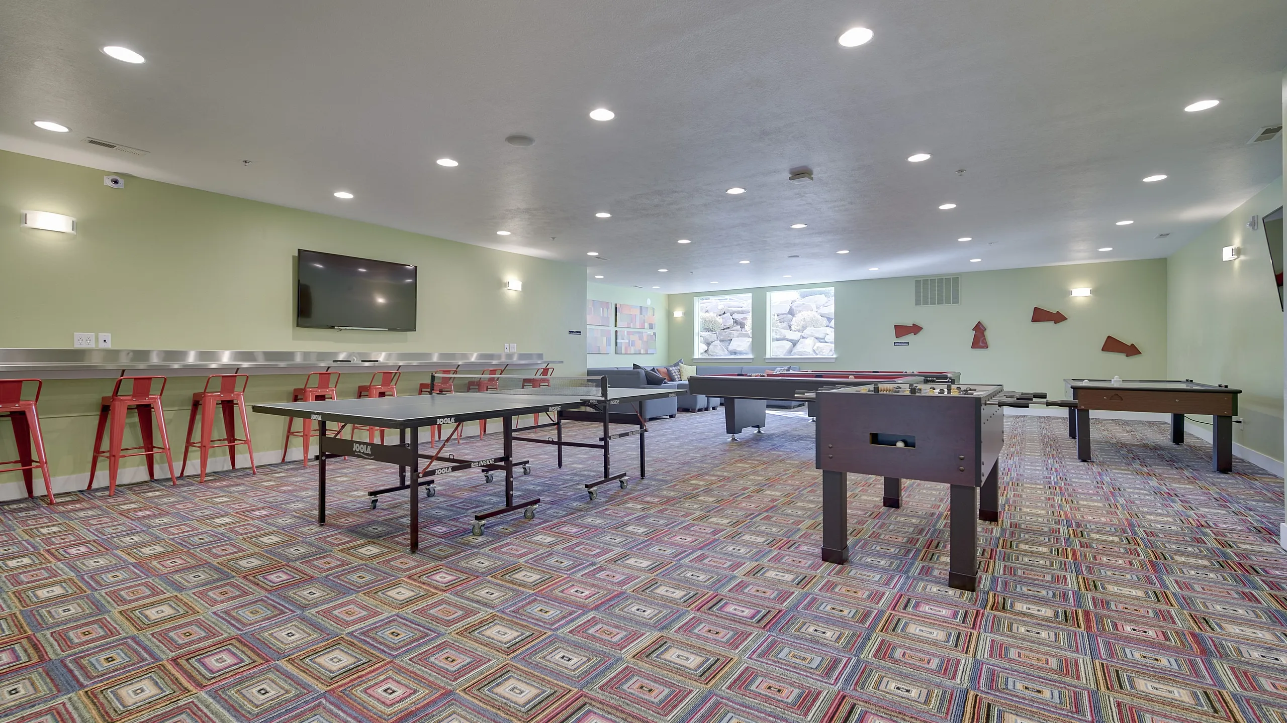 Vibrant recreation room featuring colorful patterned carpet, multiple game tables, and stylish bar seating under modern lighting.