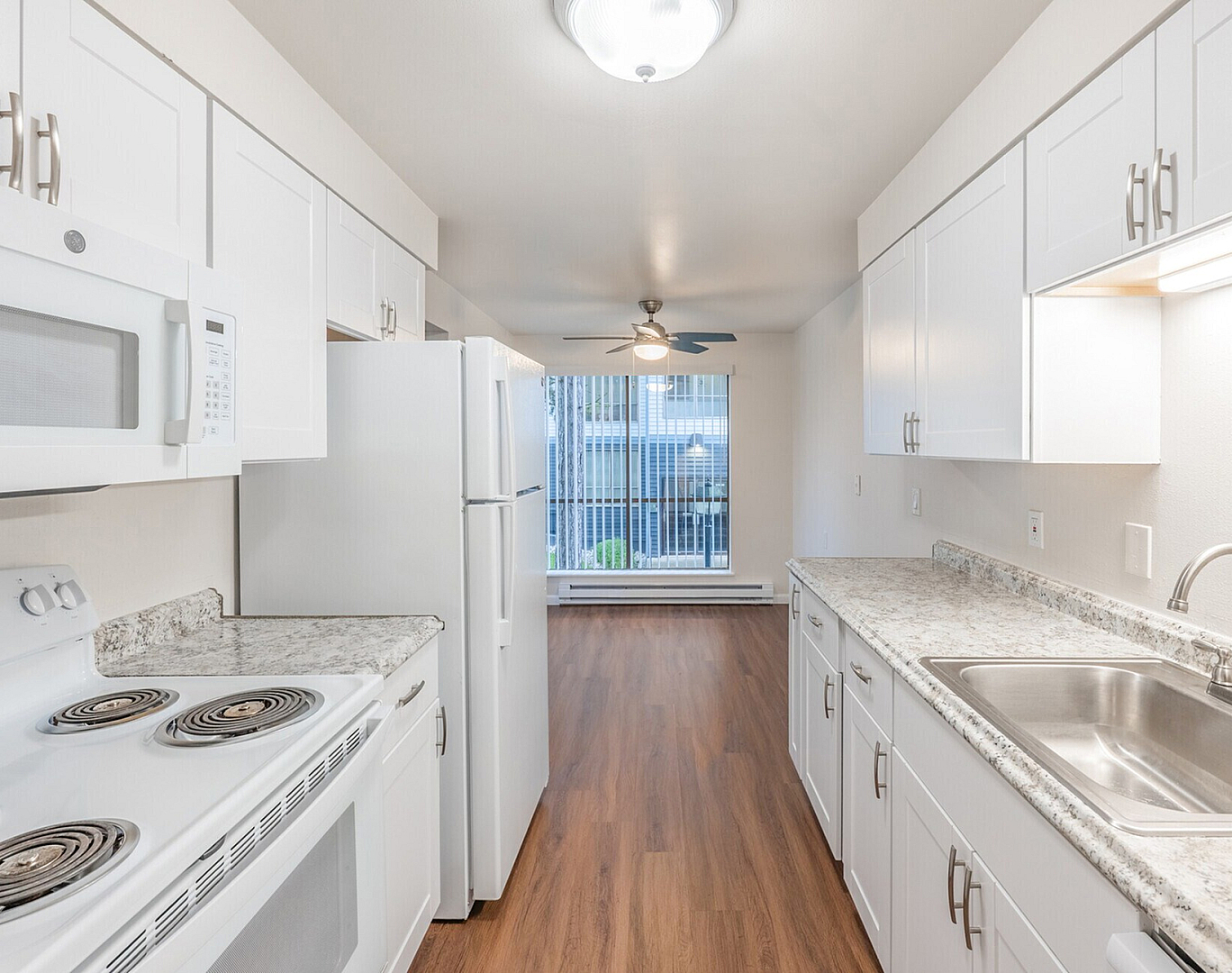 Modern kitchen with sleek white cabinetry, granite countertops, and hardwood flooring, leading to a bright living area with large windows.