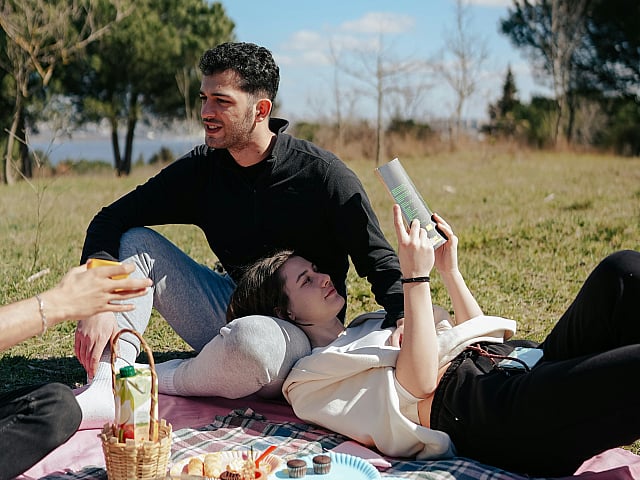 Serene outdoor picnic with people relaxing on a blanket, surrounded by snacks, fruits, and drinks under a clear blue sky.