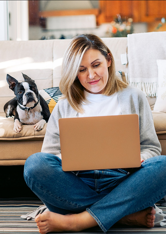 Cozy living room portrait of a woman happily working on a laptop, accompanied by a cute dog on a comfortable beige sofa, against a backdrop of soft pillows and warm decor.