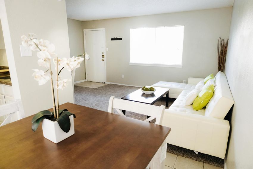 A cozy living space featuring a white sectional sofa with green accent pillows, a simple coffee table, and a dining area with a wooden table and a potted orchid. The room has neutral walls, a large window allowing natural light, and soft carpeting, creating a warm and inviting atmosphere.