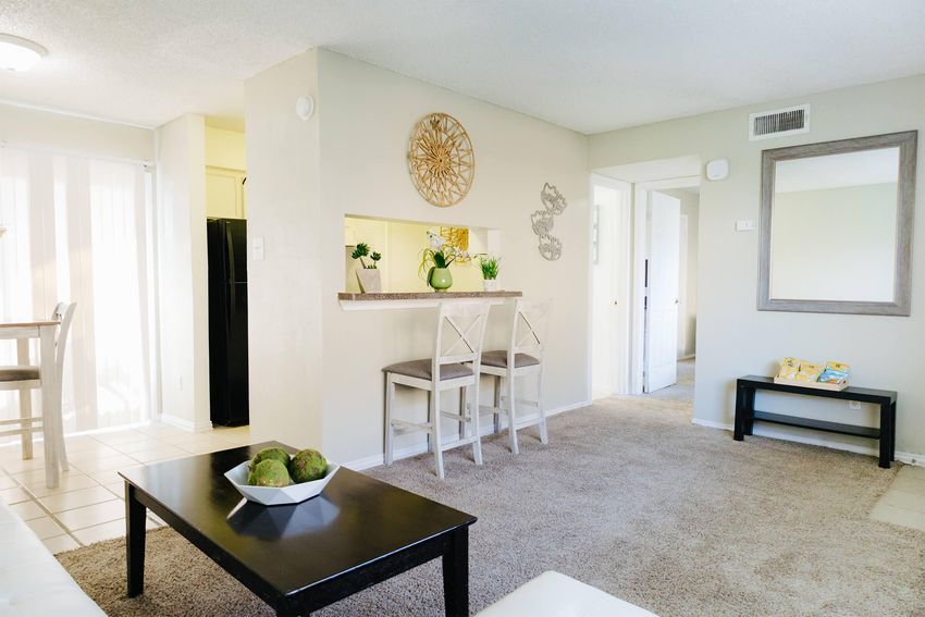 A bright and cozy living space featuring a light-colored wall with decorative accents, a bar-style table with chairs, and a mirror. The room has a plush carpet, a dark coffee table with a bowl of green decorative items, and a glimpse of a kitchen area in the background. Natural light streams in through the window.