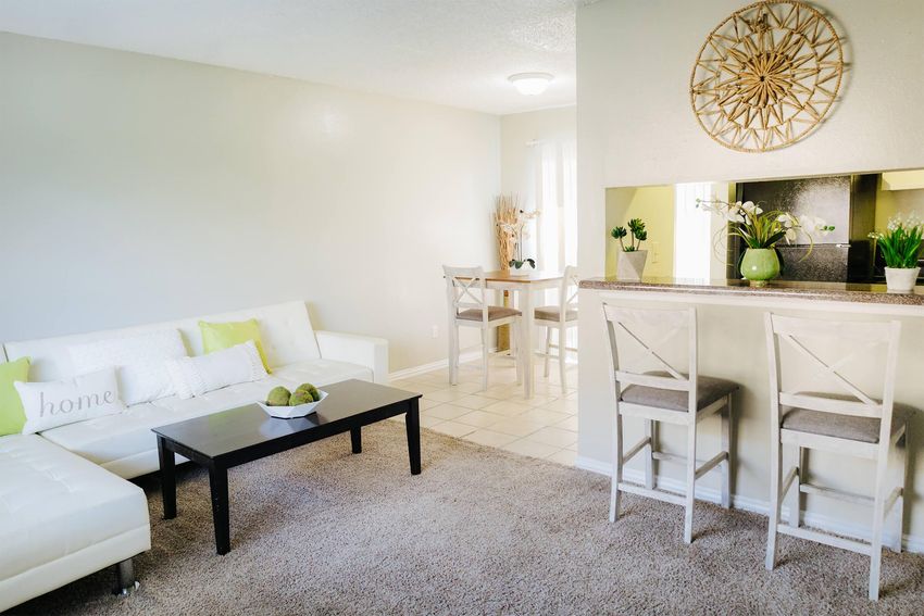 A modern living room with a white sectional sofa and a black coffee table. There are decorative pillows on the sofa, a beige carpet, and a bar area with two stools. In the background, there's a dining table with chairs and plants, and a kitchen area is visible through an opening in the wall.
